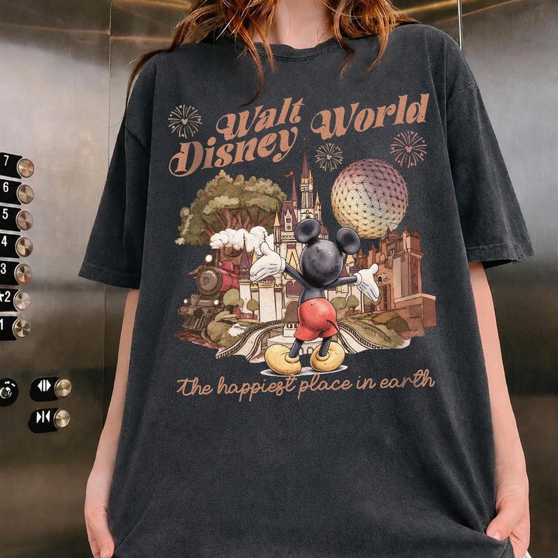 May include: Dark gray t-shirt featuring a vintage-style graphic of Mickey Mouse in front of a Disney World scene. The text "Walt Disney World" and "The happiest place in earth" are printed above and below the graphic.