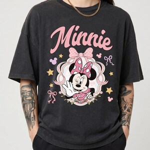 May include: A charcoal grey short-sleeve t-shirt featuring a graphic of Minnie Mouse in a pink polka-dot bow and dress. The design includes the text "Minnie" in pink script, a decorative frame, pink ribbons, and yellow stars on the front chest.
