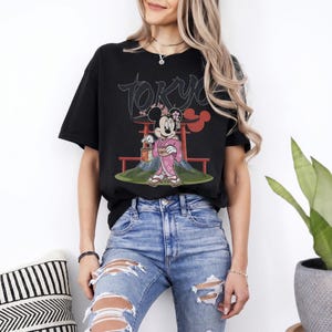 Peut inclure: T-shirt noir avec un motif de Minnie Mouse en kimono rose, tenant une lanterne, avec le mot "Tokyo" au-dessus. Le motif comprend un torii rouge et le Mont Fuji. Le t-shirt est port&eacute; avec un jean bleu.