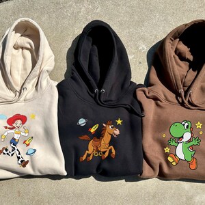 May include: Three hooded sweatshirts in beige, black, and brown. Each features a colourful cartoon character: Jessie from Toy Story, a horse with a rocket, and Yoshi from Super Mario. The sweatshirts have drawstrings.
