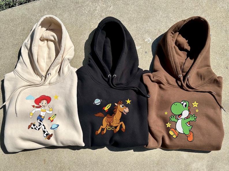 May include: Three hooded sweatshirts in beige, black, and brown. Each features a colourful cartoon character: Jessie from Toy Story, a horse with a rocket, and Yoshi from Super Mario. The sweatshirts have drawstrings.