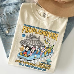May include: Cream-colored t-shirt featuring a graphic of the PeopleMover ride with Mickey, Minnie, Donald, Daisy, and Goofy. The shirt has the text "Tomorrowland Transit Authority PeopleMover" and "Keep Moving Forward To A New Tomorrow Since 1975."