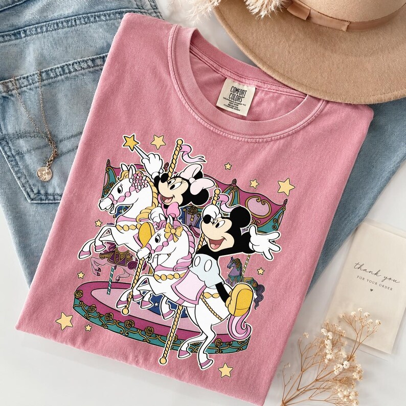 Puede incluir: Camiseta rosa con un dise&ntilde;o de carrusel con los personajes de Mickey y Minnie Mouse. El dise&ntilde;o incluye caballos blancos, estrellas y una estructura de carrusel. La camiseta se muestra con vaqueros azules, un collar y un sombrero.