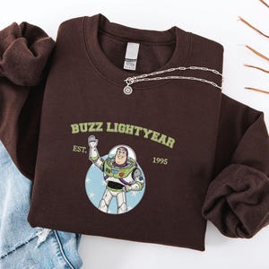 Puede incluir: Una sudadera marr&oacute;n oscuro con un gr&aacute;fico de Buzz Lightyear y el texto "Buzz Lightyear" y "EST. 1995". Un collar plateado con un colgante circular est&aacute; sobre la sudadera. La sudadera est&aacute; doblada sobre una superficie blanca.