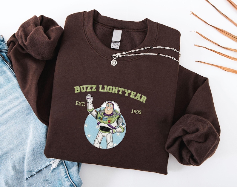 Puede incluir: Una sudadera marr&oacute;n oscuro con un gr&aacute;fico de Buzz Lightyear y el texto "Buzz Lightyear" y "EST. 1995". Un collar plateado con un colgante circular est&aacute; sobre la sudadera. La sudadera est&aacute; doblada sobre una superficie blanca.