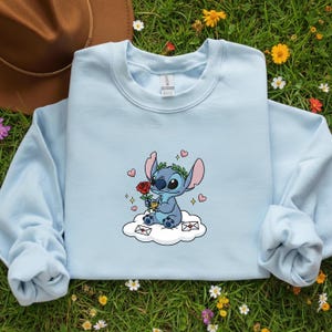Puede incluir: Sudadera azul claro con un personaje de dibujos animados de Stitch sosteniendo una rosa roja y sentado en una nube. El dise&ntilde;o incluye corazones, una corona de laurel y peque&ntilde;os sobres. La sudadera tiene mangas largas y cuello redondo.