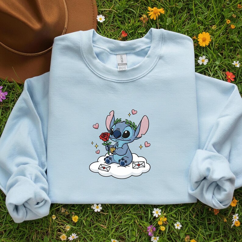 Puede incluir: Sudadera azul claro con un personaje de dibujos animados de Stitch sosteniendo una rosa roja y sentado en una nube. El dise&ntilde;o incluye corazones, una corona de laurel y peque&ntilde;os sobres. La sudadera tiene mangas largas y cuello redondo.