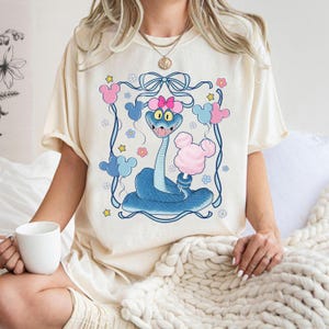 May include: Cream-colored t-shirt featuring a cartoon blue snake with a pink bow, holding cotton candy. The design includes Mickey Mouse-shaped balloons and floral accents within a blue frame. The shirt has short sleeves.
