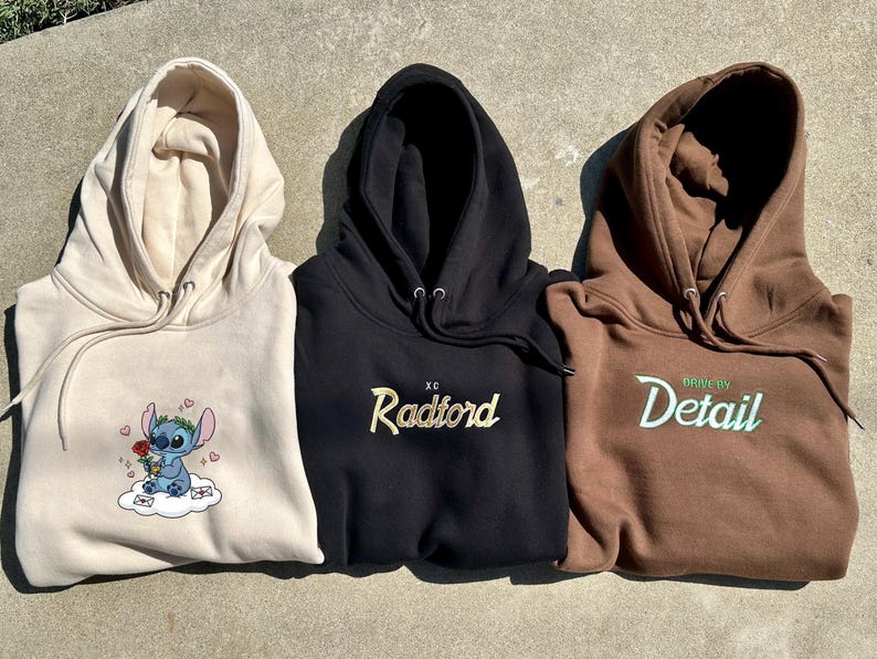 Puede incluir: Tres sudaderas con capucha en beige, negro y marr&oacute;n. La sudadera con capucha beige presenta un personaje de dibujos animados bordado. La sudadera con capucha negra tiene "Radford" bordado en dorado. La sudadera con capucha marr&oacute;n tiene "Drive By Detail" bordado en verde.