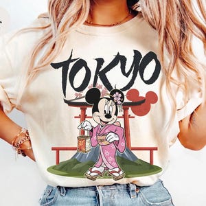 Peut inclure: T-shirt ivoire avec un motif de Minnie Mouse en kimono rose, tenant une lanterne, avec le mot "TOKYO" au-dessus. Le motif comprend une porte torii rouge et une montagne.