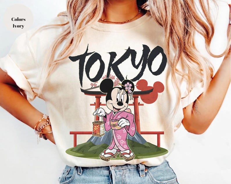 Peut inclure: T-shirt ivoire avec un motif de Minnie Mouse en kimono rose, tenant une lanterne, avec le mot "TOKYO" au-dessus. Le motif comprend une porte torii rouge et une montagne.