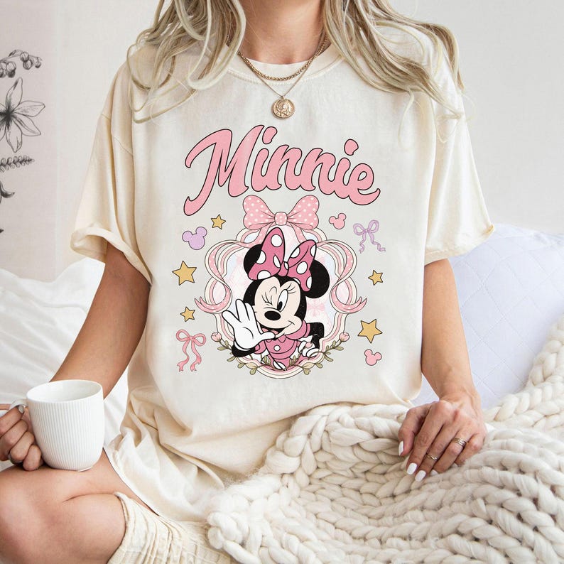 May include: A cream-colored t-shirt featuring a graphic of Minnie Mouse in a pink polka-dot bow and dress. The design includes the text Minnie in pink script, surrounded by pink ribbons, small yellow stars, and tiny pink mouse-ear silhouettes.