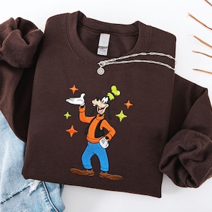 Puede incluir: Una sudadera marr&oacute;n oscuro con una imagen bordada de Goofy, con un chaleco naranja, pantalones azules y un sombrero verde y amarillo. El dise&ntilde;o incluye acentos de estrellas. Un collar plateado con un colgante est&aacute; drapeado sobre la sudadera.