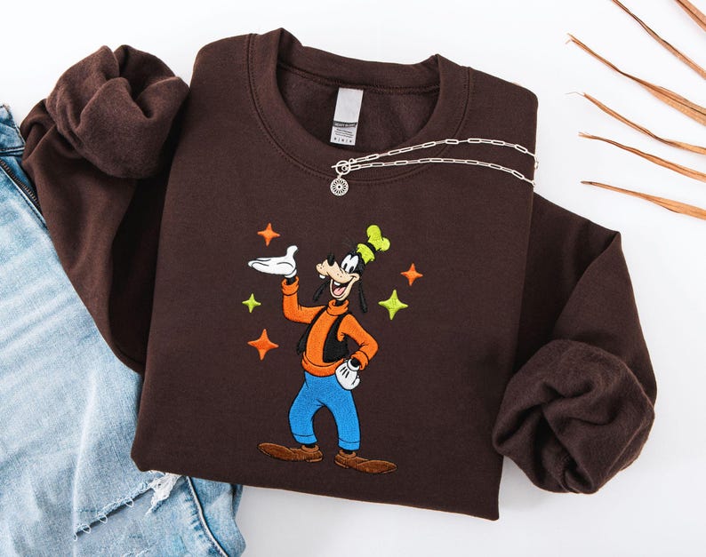 Puede incluir: Una sudadera marr&oacute;n oscuro con una imagen bordada de Goofy, con un chaleco naranja, pantalones azules y un sombrero verde y amarillo. El dise&ntilde;o incluye acentos de estrellas. Un collar plateado con un colgante est&aacute; drapeado sobre la sudadera.