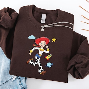 May include: A brown sweatshirt with an embroidered cartoon character in a red hat and cow-print trousers. The design includes a yellow star, a rocket, and a planet. A silver necklace with a pendant is draped across the sweatshirt.