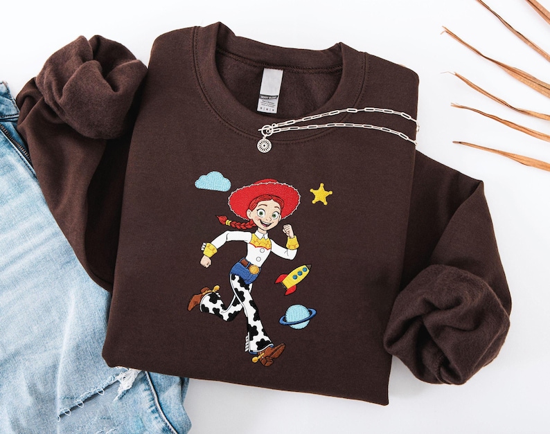 May include: A brown sweatshirt with an embroidered cartoon character in a red hat and cow-print trousers. The design includes a yellow star, a rocket, and a planet. A silver necklace with a pendant is draped across the sweatshirt.