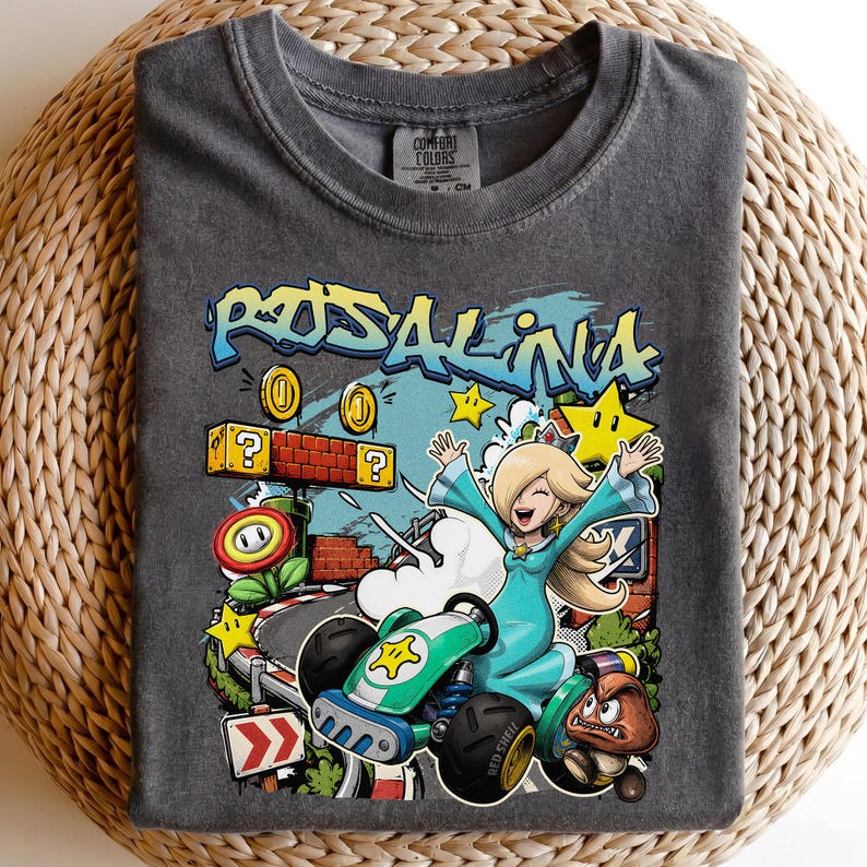 May include: Dark gray t-shirt featuring a colorful graphic of Rosalina from the Mario Kart series. The design includes Rosalina in a kart, coins, stars, and the text "Rosalina". The shirt is made of a soft material.