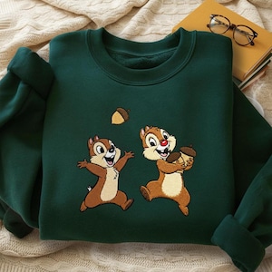 Könnte beinhalten: Dunkelgrünes Sweatshirt mit gestickten Comic-Chipmunks, einer mit einer Eichel und der andere mit ausgestreckten Armen. Eine beige Eichel schwebt über ihnen. Das Sweatshirt liegt auf einer cremefarbenen Decke.