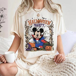 May include: A cream-colored t-shirt featuring a graphic of Mickey Mouse dressed as a vampire. The design includes the text "HALLOWEEN PARTY," pumpkins, a witch hat, a skull, a cauldron, and bare tree branches framed by vines and bats.