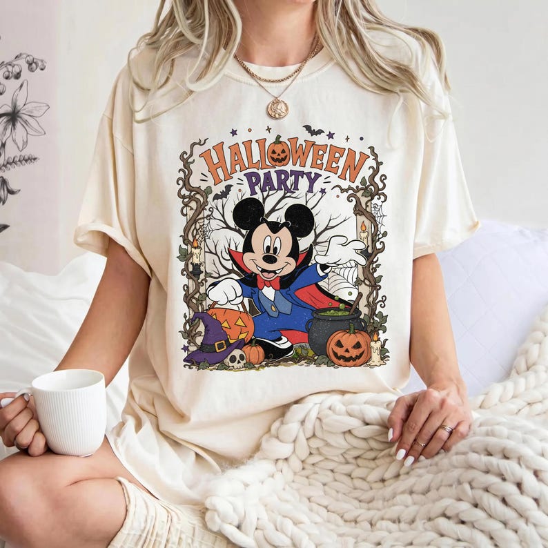 May include: A cream-colored t-shirt featuring a graphic of Mickey Mouse dressed as a vampire. The design includes the text "HALLOWEEN PARTY," pumpkins, a witch hat, a skull, a cauldron, and bare tree branches framed by vines and bats.