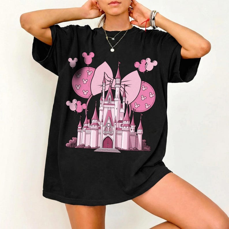 May include: Black t-shirt featuring a pink castle design with a large bow and Mickey Mouse head balloons. The castle has multiple turrets and a central entrance. The design is centered on the front of the shirt.