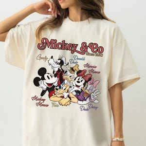 May include: Cream-colored t-shirt featuring a graphic of Mickey Mouse and friends. The design includes Mickey, Minnie, Donald, Daisy, Goofy, and Pluto, with the text "Mickey & Co Since 1928".