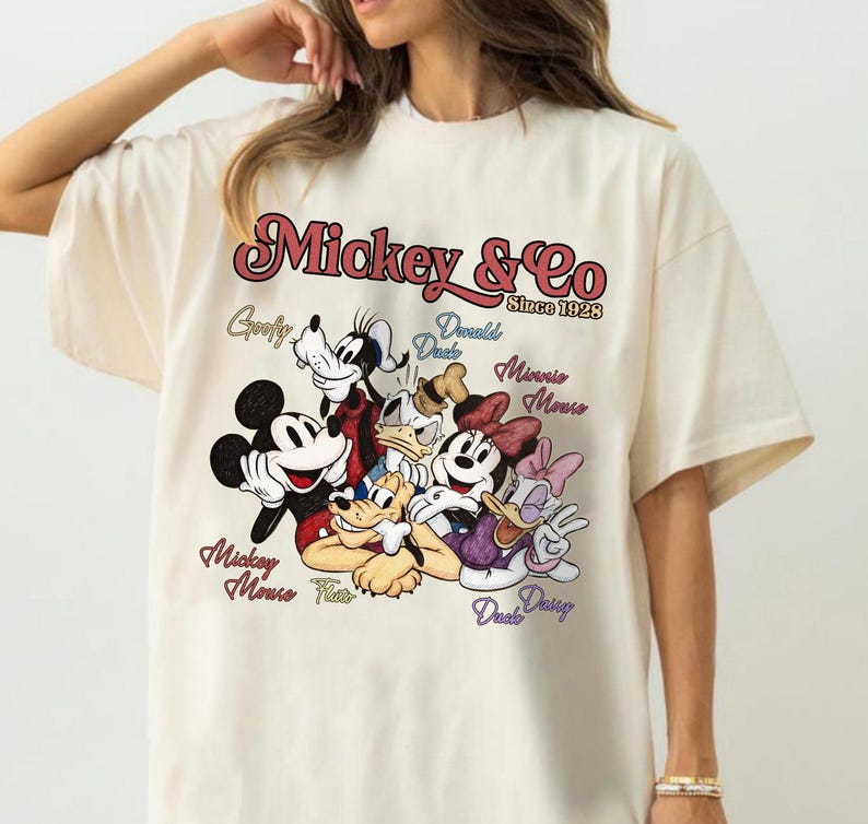 May include: Cream-colored t-shirt featuring a graphic of Mickey Mouse and friends. The design includes Mickey, Minnie, Donald, Daisy, Goofy, and Pluto, with the text "Mickey & Co Since 1928".
