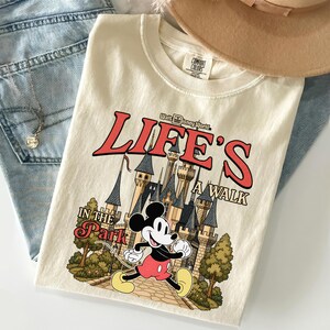 Puede incluir: Camiseta color crema con un gr&aacute;fico de Mickey Mouse corriendo frente a un castillo, con el texto "LIFE'S A WALK IN THE PARK" en rojo y negro. La camiseta est&aacute; doblada sobre unos vaqueros azules y un sombrero marr&oacute;n.