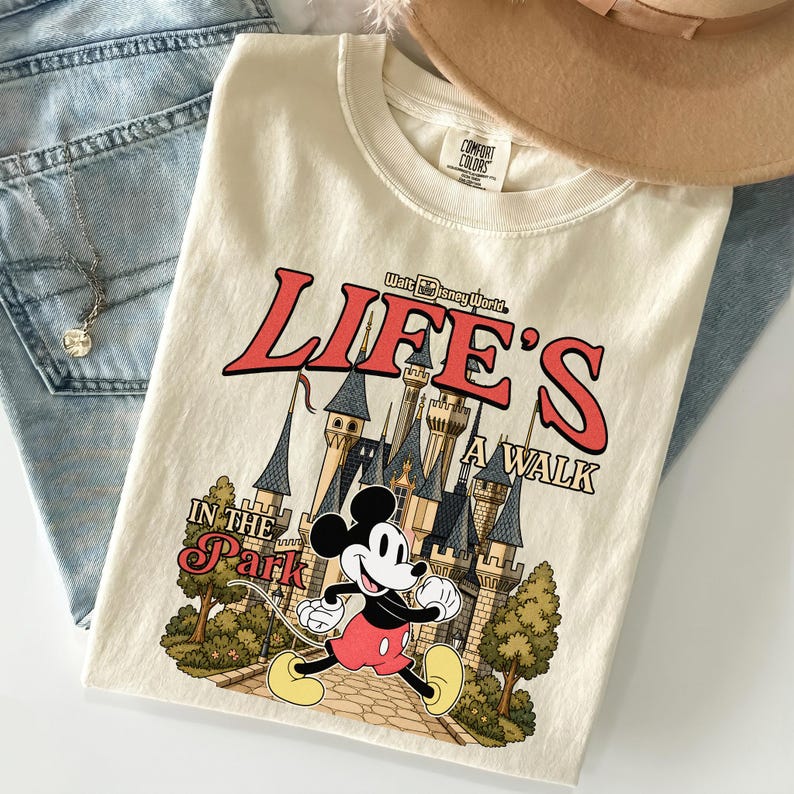 Puede incluir: Camiseta color crema con un gr&aacute;fico de Mickey Mouse corriendo frente a un castillo, con el texto "LIFE'S A WALK IN THE PARK" en rojo y negro. La camiseta est&aacute; doblada sobre unos vaqueros azules y un sombrero marr&oacute;n.