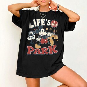 Puede incluir: Una camiseta negra de manga corta con un gr&aacute;fico de Mickey Mouse caminando. El texto dice "LIFE'S A WALK IN THE PARK" junto a una ilustraci&oacute;n de un castillo, estrellas y una se&ntilde;al. El dise&ntilde;o utiliza colores rojo, blanco, amarillo y azul.