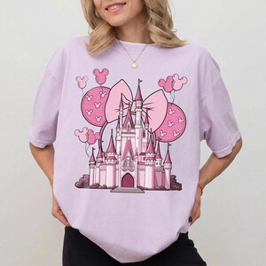 May include: A light purple t-shirt featuring a pink castle design with a large bow and Mickey Mouse ear balloons. The castle has multiple turrets and a central entrance. The design is in a cartoon style.