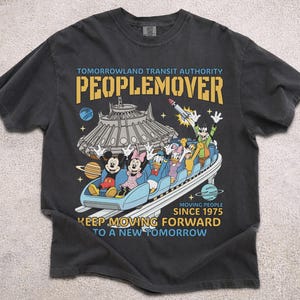May include: Dark gray t-shirt featuring a colorful graphic of the PeopleMover ride with Mickey Mouse, Minnie Mouse, Donald Duck, Daisy Duck, and Goofy. The text reads "Tomorrowland Transit Authority PeopleMover" and "Keep Moving Forward to a New Tomorrow."