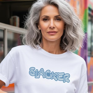May include: White t-shirt with the word "SLACKER" in light blue bubble letters. The shirt is being worn by a person with gray hair, standing in front of a colorful food truck.