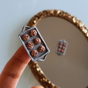 May include: A miniature baking tray filled with chocolate chip cookies, held in a person's hand in front of a gold-framed mirror.