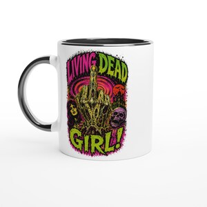 May include: White ceramic mug with a black rim and handle. The mug features a graphic design with the words "LIVING DEAD GIRL!" in green and pink, along with a zombie hand giving the middle finger, a skull, and a woman's face.