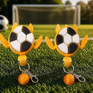 May include: Two yellow and black soccer ball keychains with orange accents. Each keychain features a soccer ball design with white and black pentagons, and a small orange ball charm. The keychains are on a green field with a soccer goal in the background.
