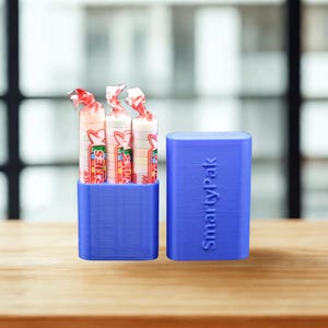 May include: A blue plastic SmartyPak container with a textured finish. The container is divided into two parts, with one section holding three rolls of Smarties candy. The word SmartyPak is embossed vertically on the side of the container.