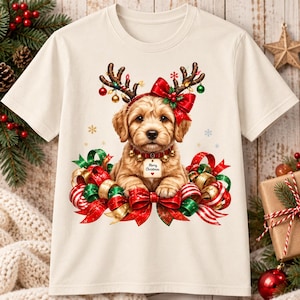 Könnte beinhalten: Ein cremefarbenes T-Shirt mit einer Grafik eines goldenen Welpen, der ein Rentiergeweih mit bunten Ornamenten und einer roten Schleife trägt. Der Welpe hat ein Halsband mit einem Schild, auf dem Merry Christmas steht, und ist von roten und grünen Bändern umgeben.
