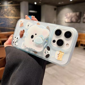 May include: A clear phone case with a light blue tint, adorned with cartoon dog illustrations. The case features a large, 3D white dog with a blue hat and a pop-up grip. Additional dog-themed stickers are scattered across the case.