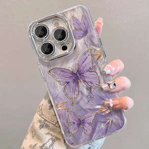 Peut inclure: Coque de téléphone transparente avec un motif de papillons violets et dorés. La coque présente trois papillons et des accents dorés. L'arrière-plan a un aspect texturé et brossé. L'objectif de la caméra est visible.