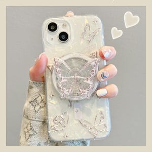 May include: Clear phone case with a pearlescent finish, featuring embossed butterflies and star accents. A silver butterfly-shaped phone grip is attached. The case has a dual-camera cutout.