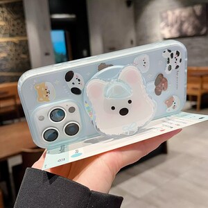 May include: A light blue phone case featuring various cartoon dog illustrations. The case has a white dog-shaped pop socket. The phone case has a clear protective layer and a camera cutout.