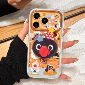 May include: A clear phone case with a cartoon penguin design. The case has a yellow background with various penguin illustrations and a 3D penguin pop-up. The case is held by a person.