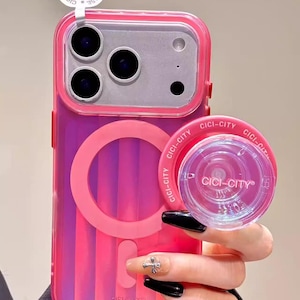 Puede incluir: Una funda de teléfono rosa y transparente con un pop socket a juego. La funda tiene un diseño a rayas y el pop socket es redondo con el texto "CICI-CITY". La funda tiene un recorte para la cámara y un anillo magnético.
