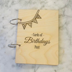 Puede incluir: Un álbum de recortes de madera con un encuadernador de anillas. La cubierta es de color marrón claro y tiene un diseño grabado con láser de una hilera de banderas con el texto "Cards of Birthdays Past".