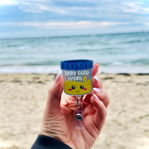 K&ouml;nnte beinhalten: Ein glitzernder Acryl-Ausweishalter in Form eines Probenbechers mit blauem Deckel, gelber Fl&uuml;ssigkeit und einem l&auml;chelnden Gesicht. Der Text "URINE GOOD HANDS" ist in wei&szlig;en Buchstaben &uuml;ber einem kleinen gelben Tropfensymbol aufgedruckt.