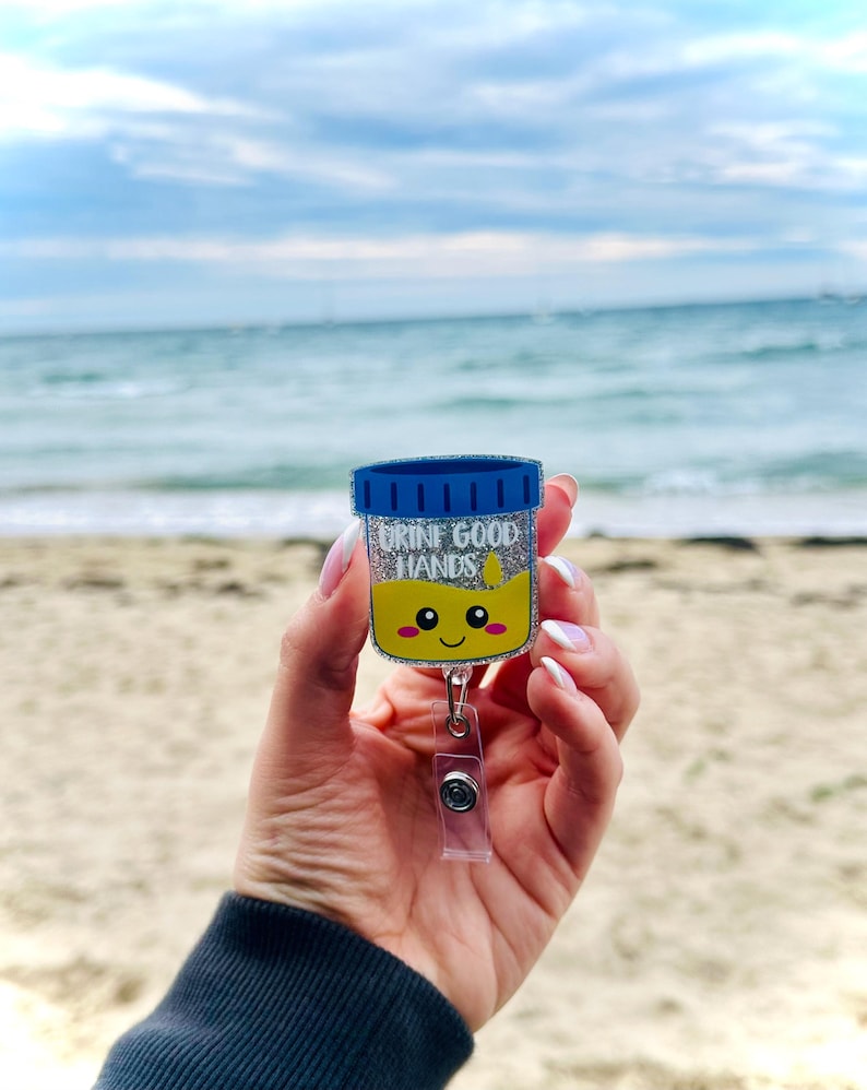 K&ouml;nnte beinhalten: Ein glitzernder Acryl-Ausweishalter in Form eines Probenbechers mit blauem Deckel, gelber Fl&uuml;ssigkeit und einem l&auml;chelnden Gesicht. Der Text "URINE GOOD HANDS" ist in wei&szlig;en Buchstaben &uuml;ber einem kleinen gelben Tropfensymbol aufgedruckt.