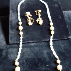 May include: A white pearl necklace with a gold clasp and matching earrings. The necklace and earrings are on a black velvet background.