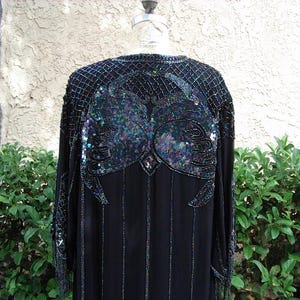 May include: Black evening gown with intricate sequin and bead embellishments. The gown features a detailed design on the shoulders and back, with vertical beaded stripes down the front. The sleeves have fringe detailing. The dress is displayed on a mannequin.