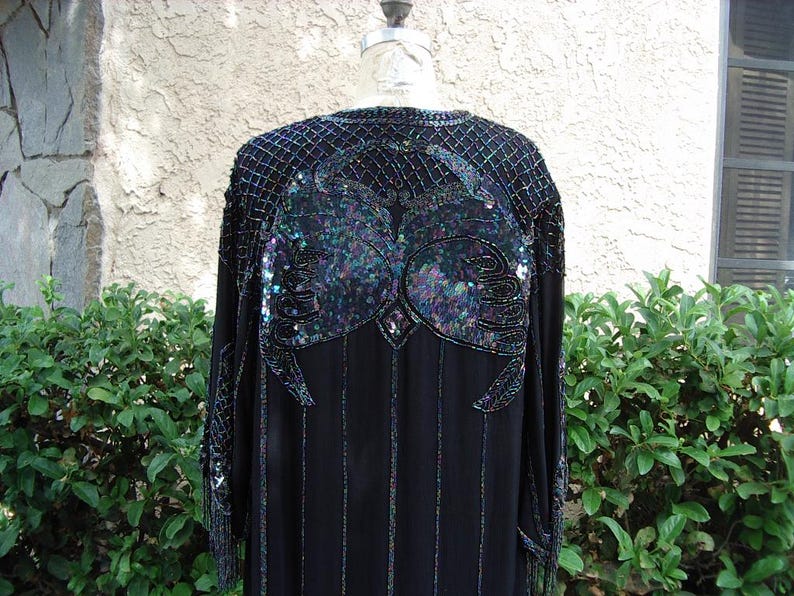 May include: Black evening gown with intricate sequin and bead embellishments. The gown features a detailed design on the shoulders and back, with vertical beaded stripes down the front. The sleeves have fringe detailing. The dress is displayed on a mannequin.